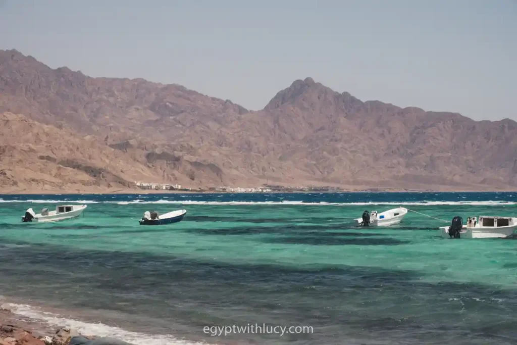 Small white boats float on the turquoise water near a mountainous background.