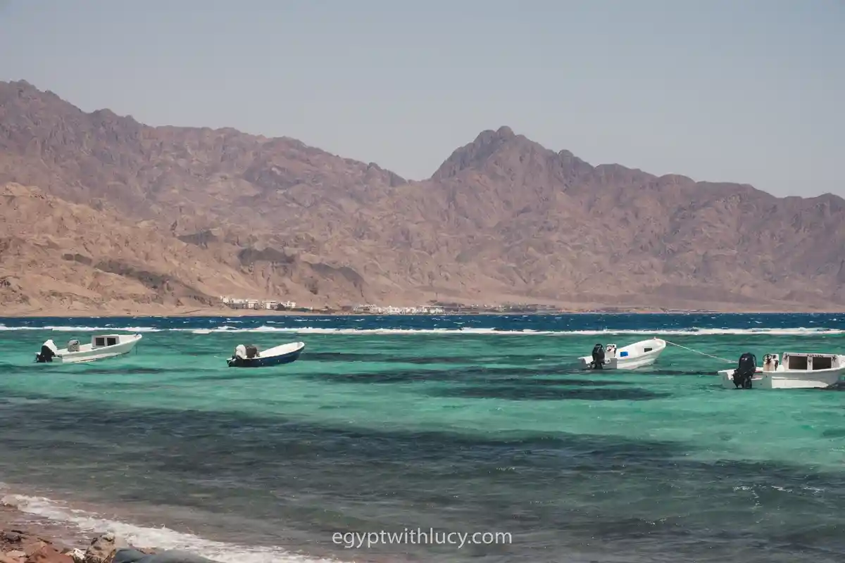 Small white boats float on the turquoise water near a mountainous background.