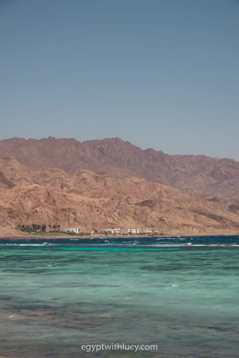 A clear coral reef layer shows through turquoise ocean water below distant desert mountains.