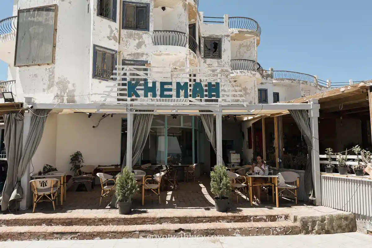 A woman sits at an outdoor table under the sign for Khemah cafe.