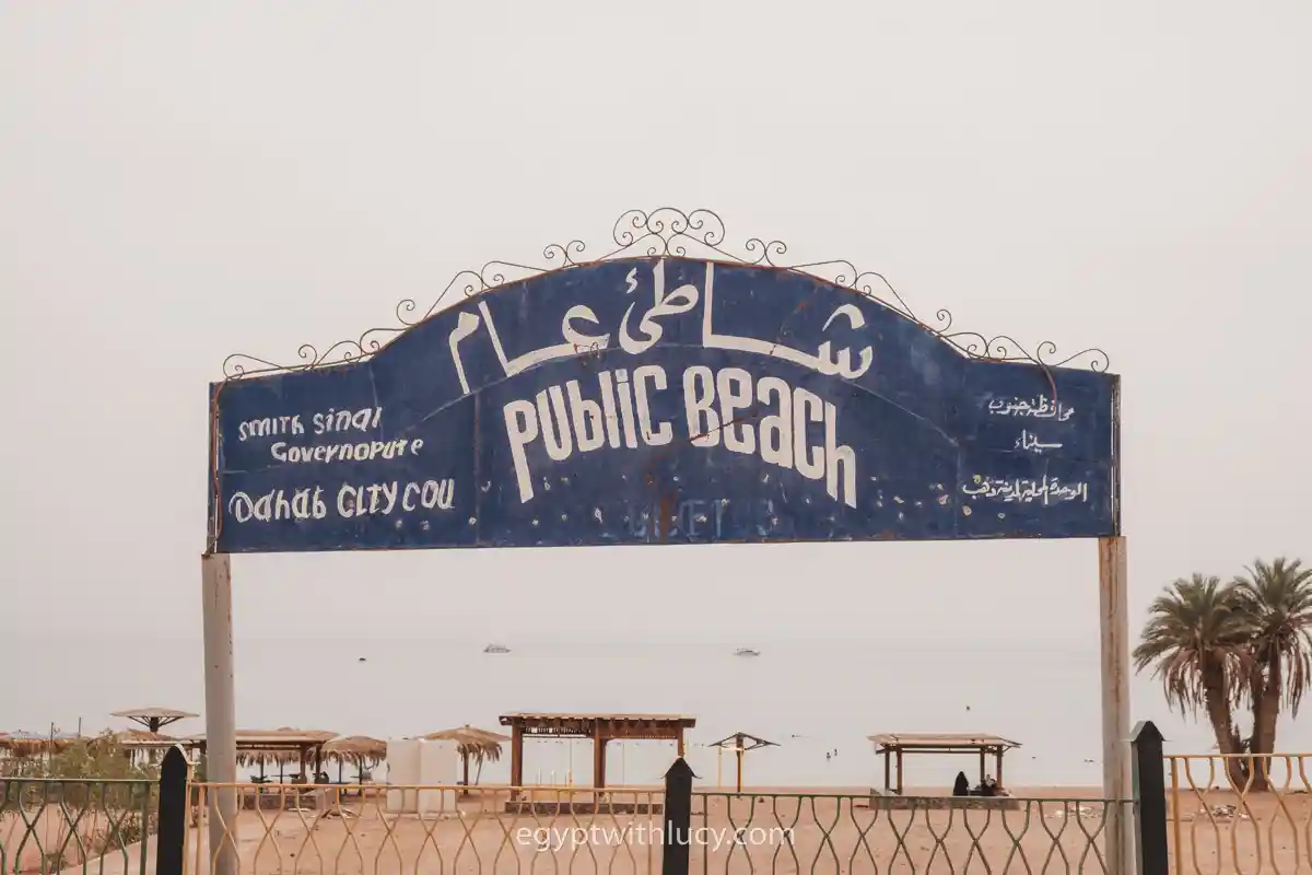 A blue archway sign with white lettering that reads Public Beach in English and Arabic stands in front of a beach.
