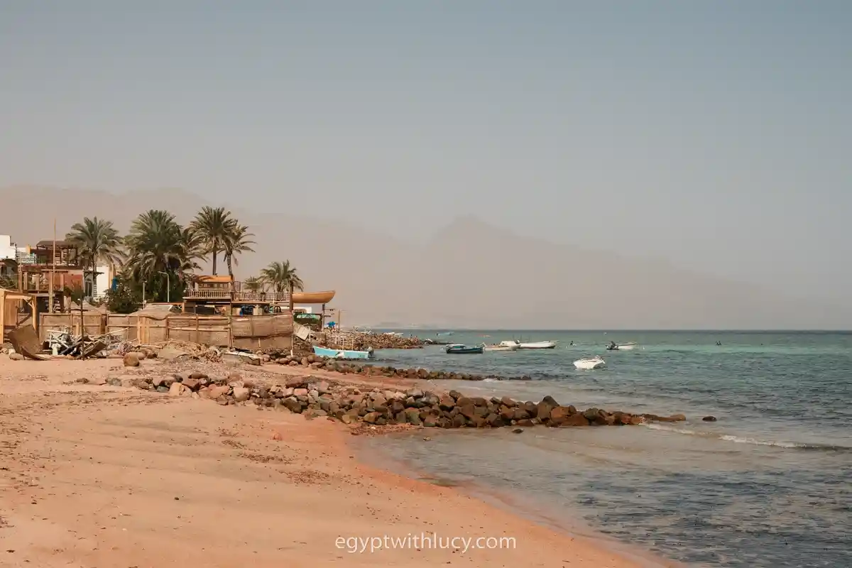 sandy-beach-dahab Small boats float on the water near Dahab's sandy coastline with palm trees.