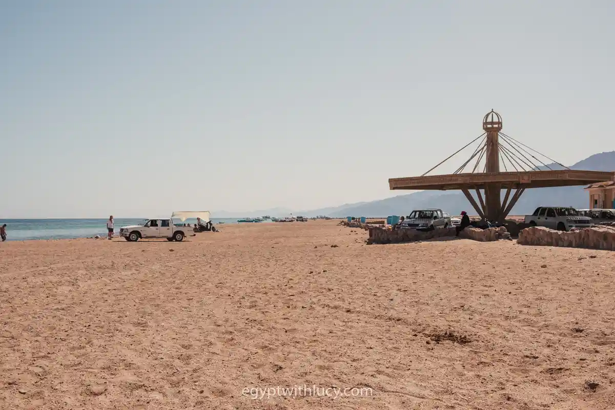 Pickup trucks park on the sand near a large wooden shade structure at The Islands Beach.