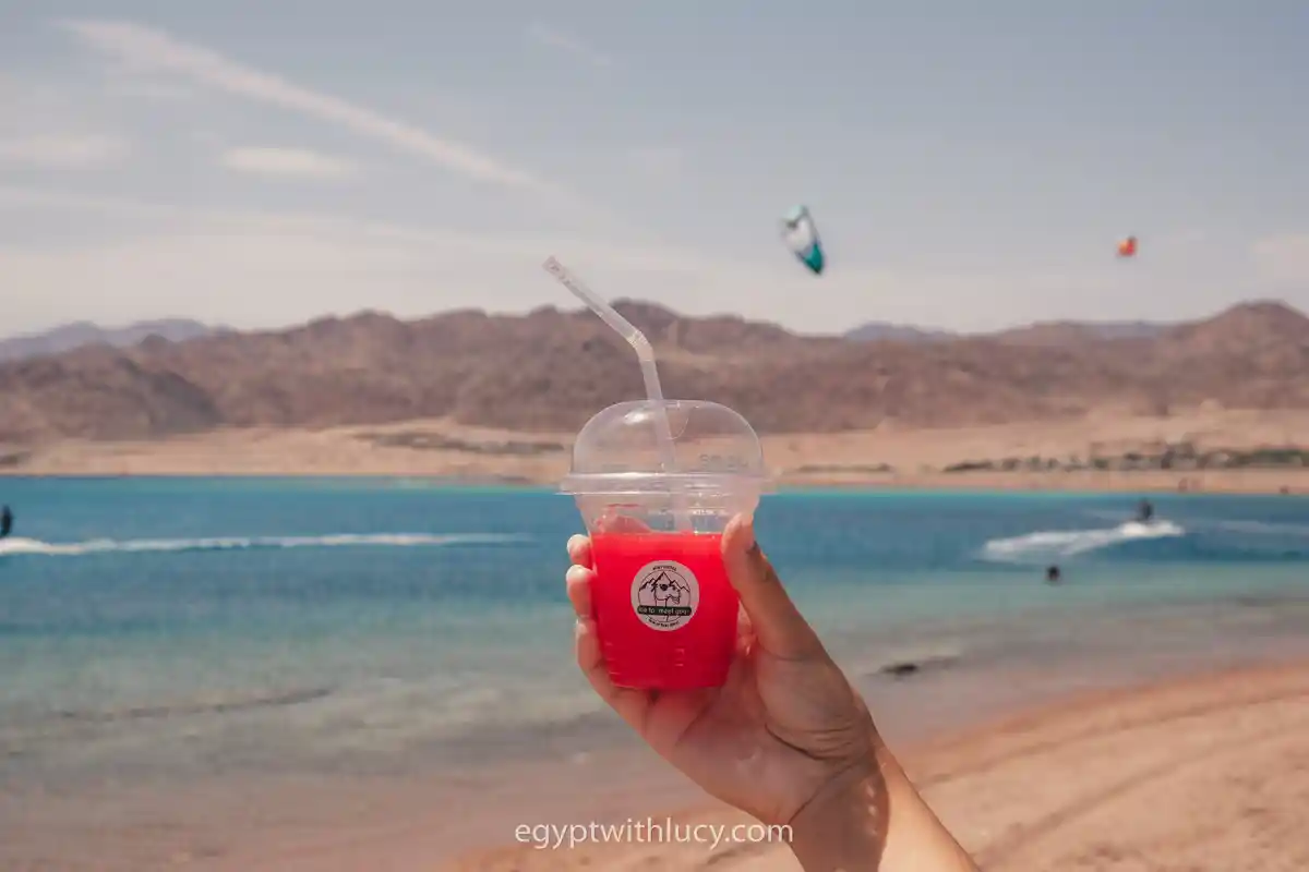 A hand holds a plastic cup of red watermelon juice in front of Laguna Beach.