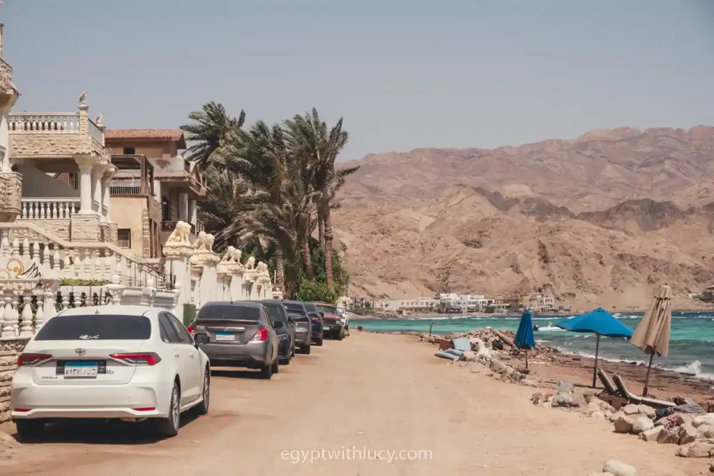 Cars parked along a coastal dirt road next to buildings and mountains in the Assala neighbourhood of Dahab.