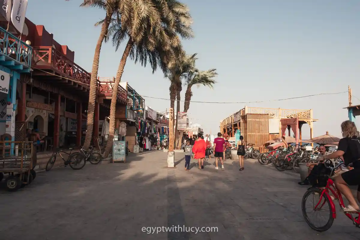 People walking down a paved pedestrian beach promenade in dahab which is lined with palm trees and cafes.