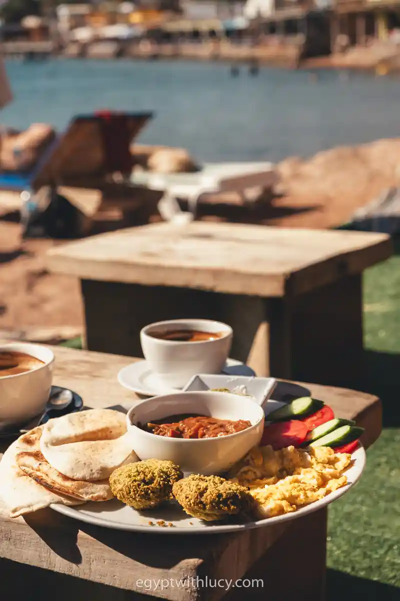An Egyptian breakfast platter with falafel, foul, scrambled eggs, bread, and salad by the sea.