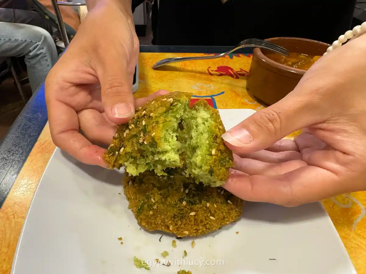 Hands breaking open a fried green tamiya patty covered in sesame seeds.