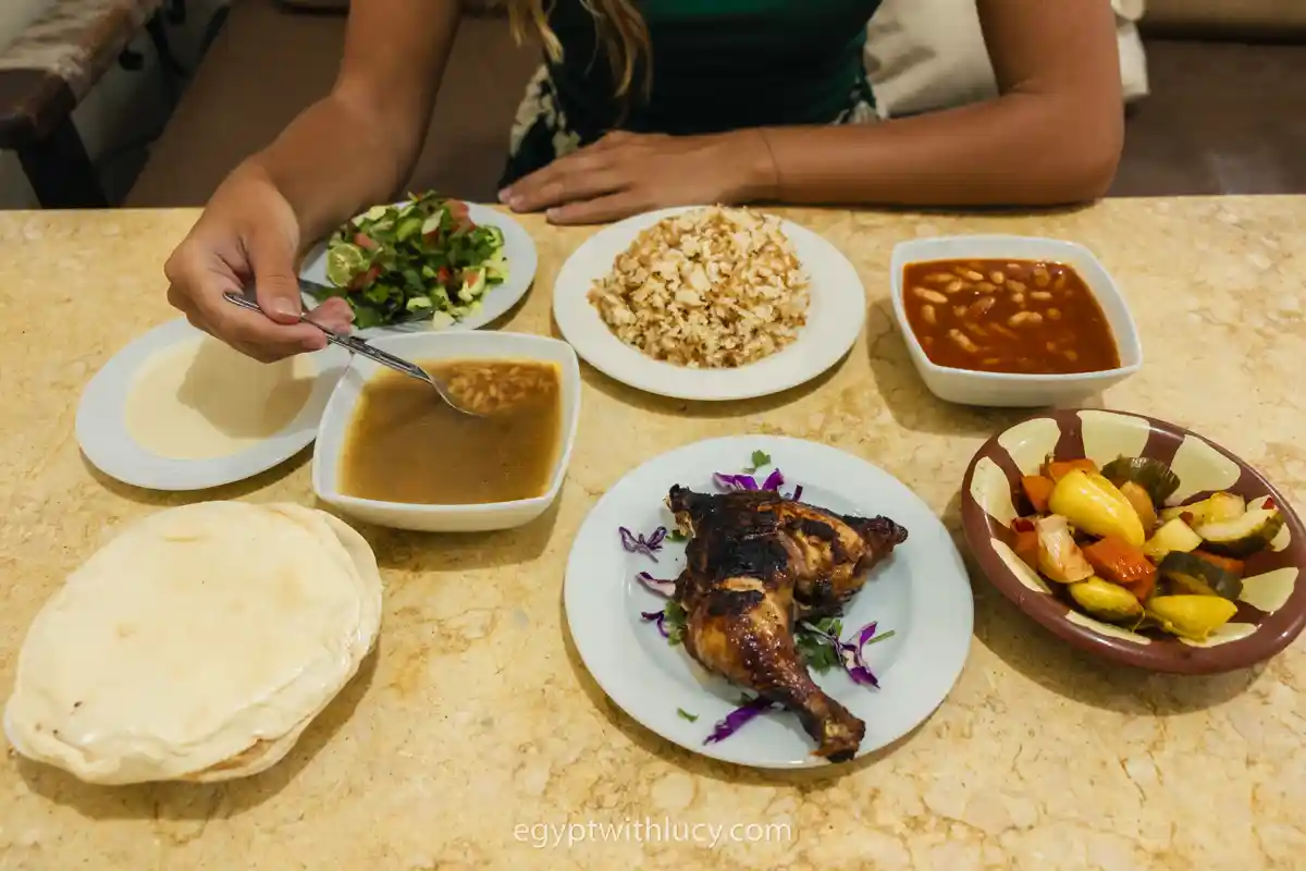 A dining table set with grilled chicken, rice, soup, pita bread, and salad bowls.