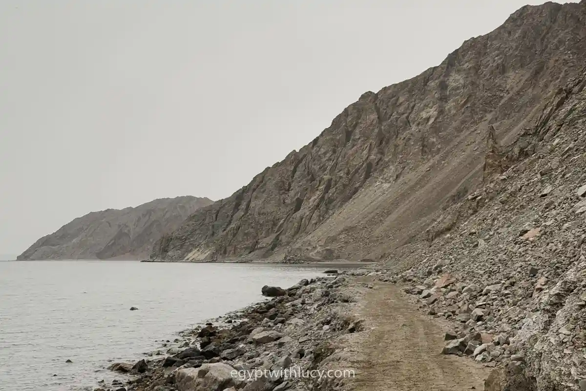 A rocky dirt path running alongside the sea beneath a steep, barren mountain.