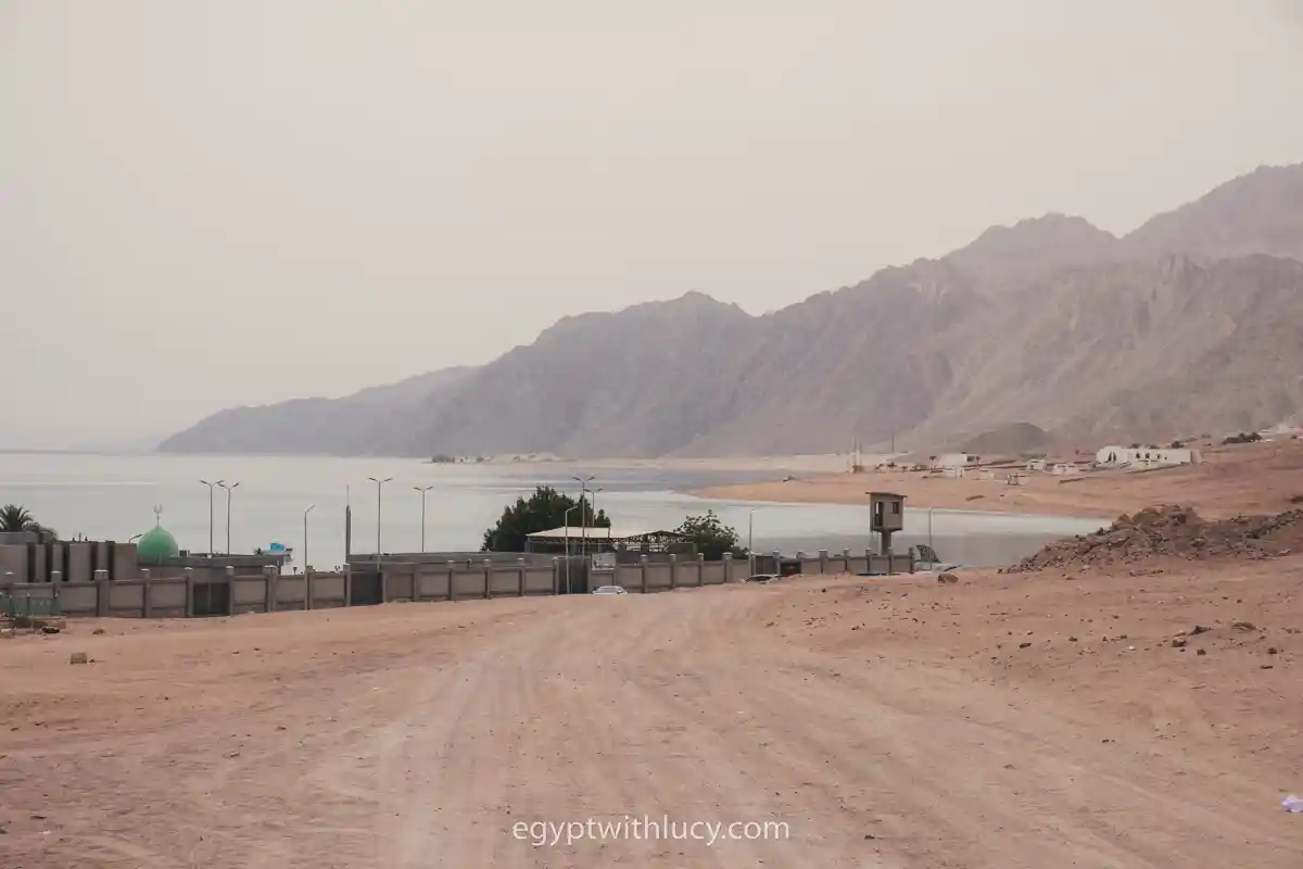 A wide dirt road leading toward a coastal area with distant mountains.