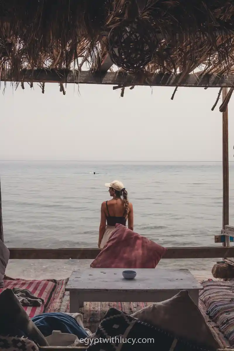 A woman in a cap standing under a thatched roof looking out at the sea.