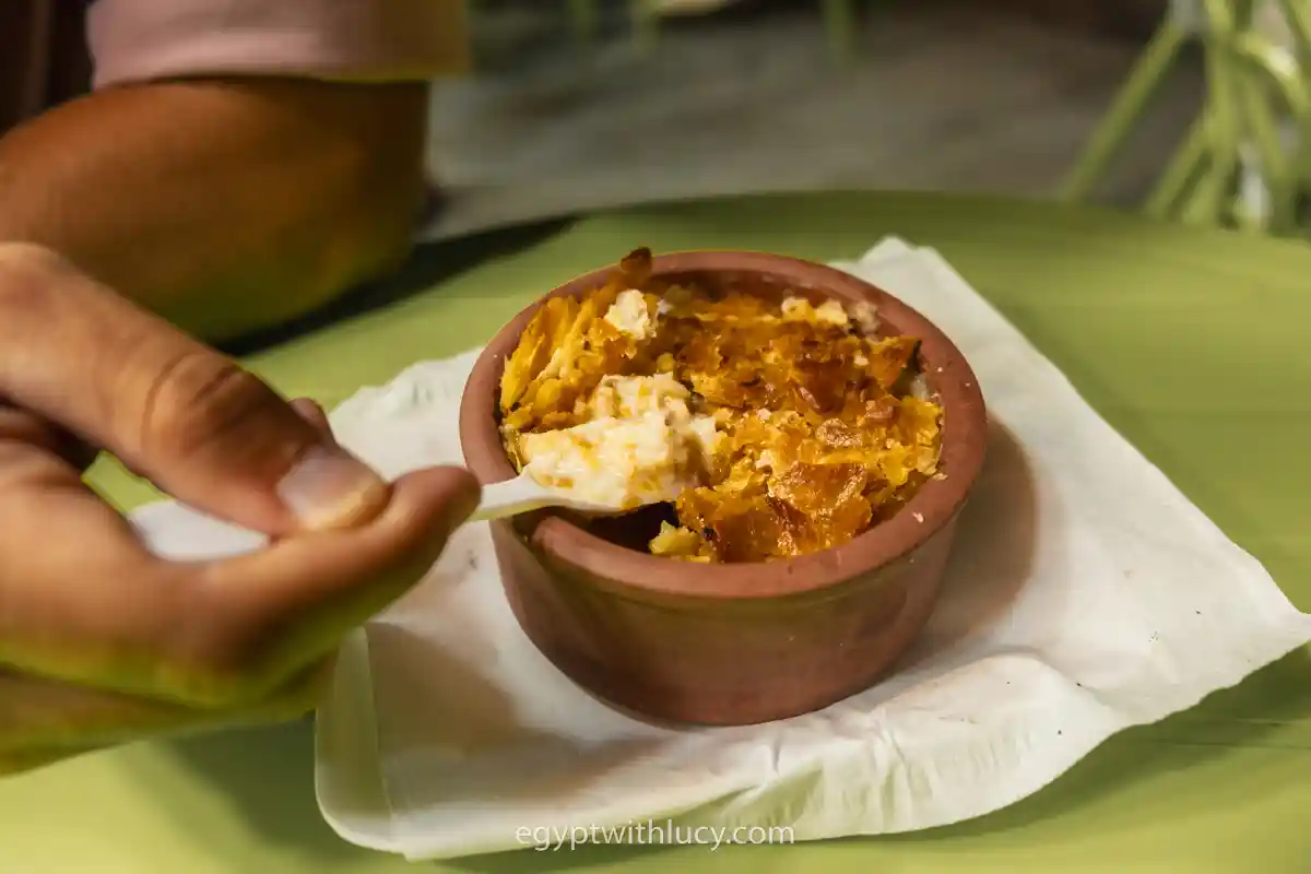 umm-ali-dessert A hand using a white plastic spoon to scoop baked Om Ali pudding from a clay pot.